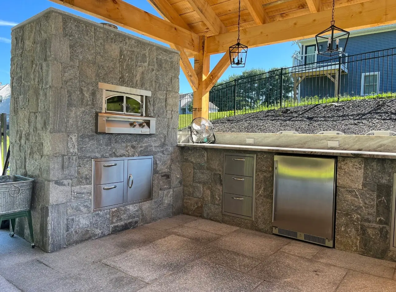 Covered outdoor kitchen with stone surround, built-in stainless grill, drawers, and an undercounter refrigerator.