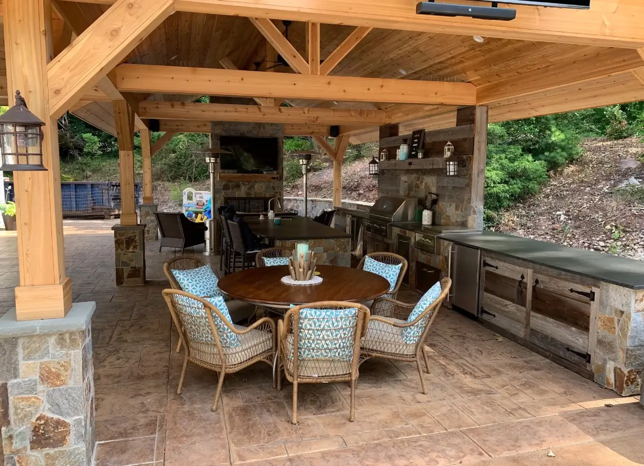 Covered patio with timber beams, dining table and chairs, and an outdoor kitchen with stone counters and stainless appliances.