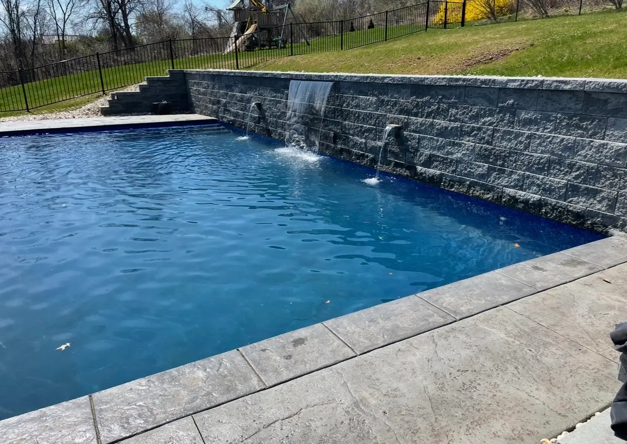 In-ground vinyl liner pool with deep-blue liner, stone coping, and waterfall features along a retaining wall.