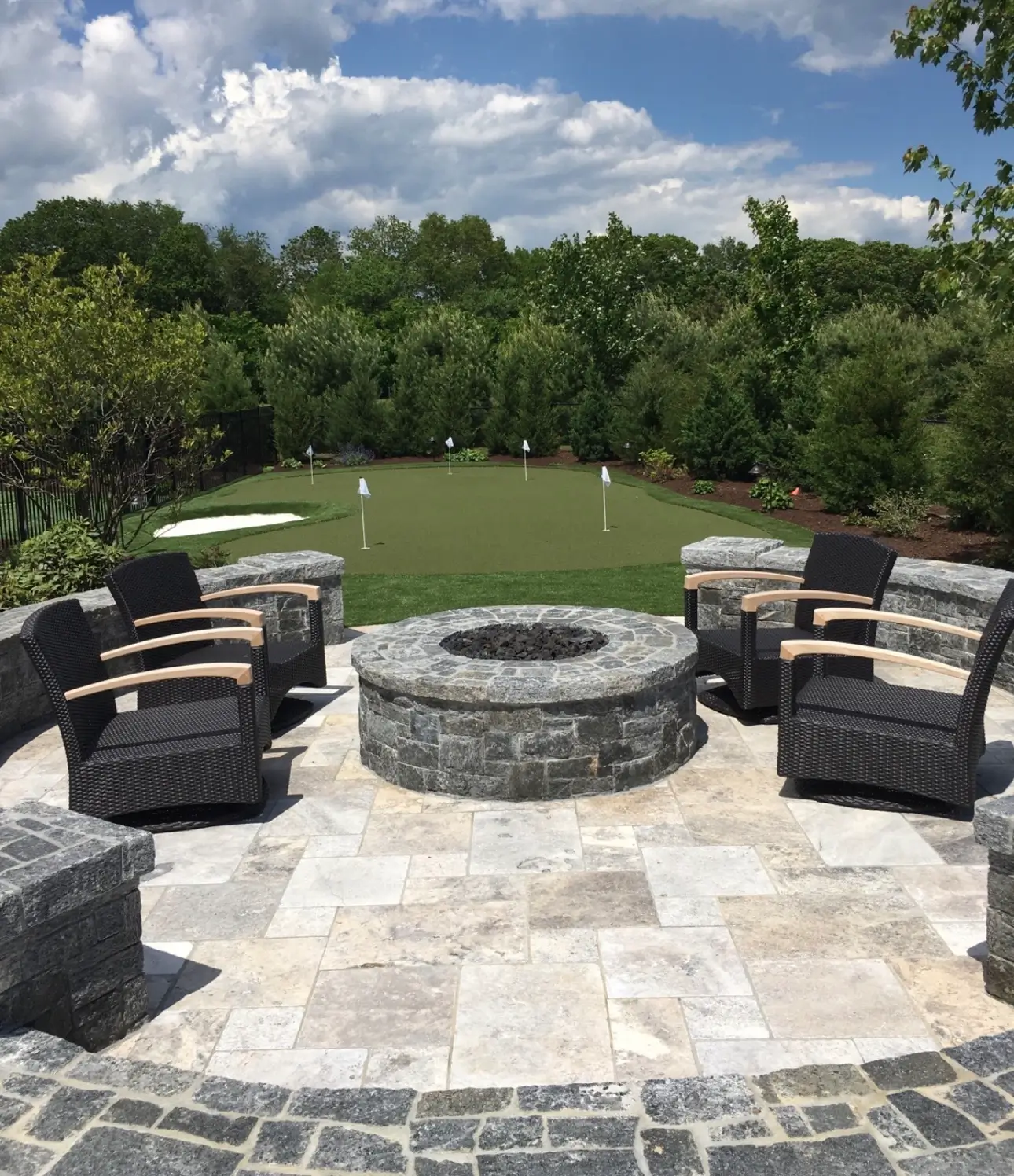Circular stone fire pit surrounded by chairs on a paver patio, with a putting green and small flags in the background.