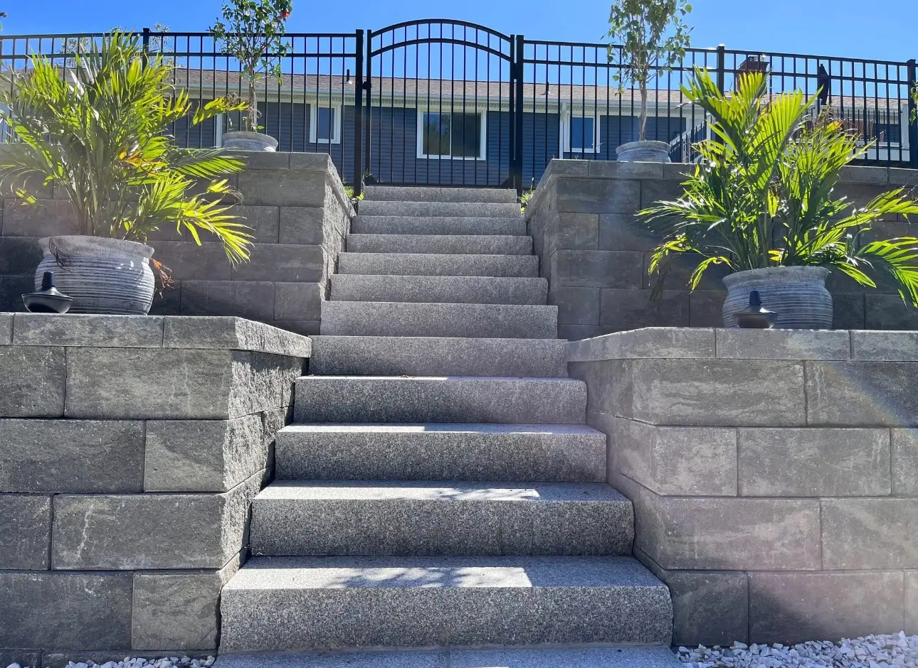 Wide granite steps between stacked-stone retaining walls with large planters and a black metal fence at the top.