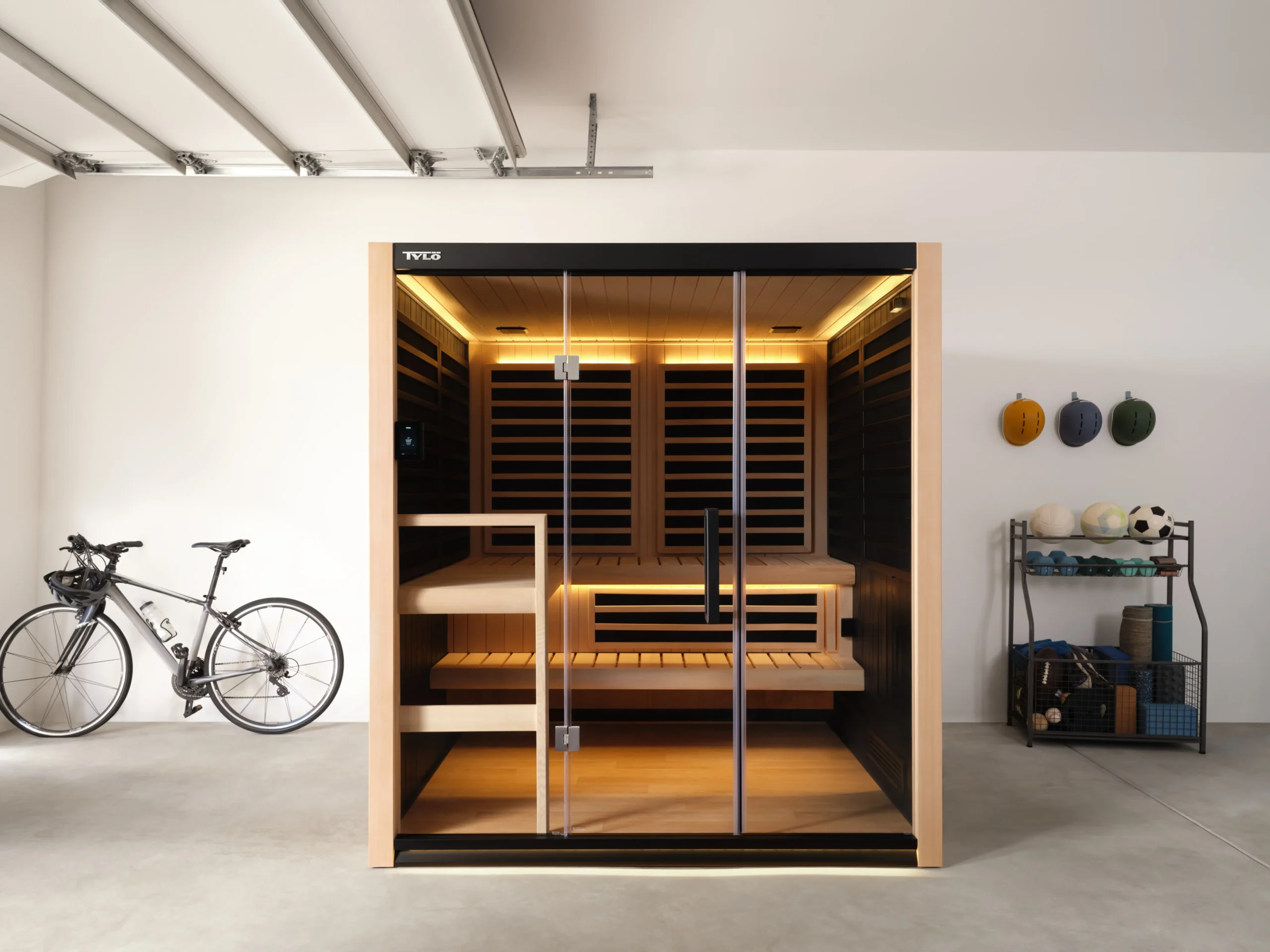 A Tylö indoor sauna cabin with a light wood finish and glass doors installed in a bright garage next to a bicycle and a rack of sports equipment.