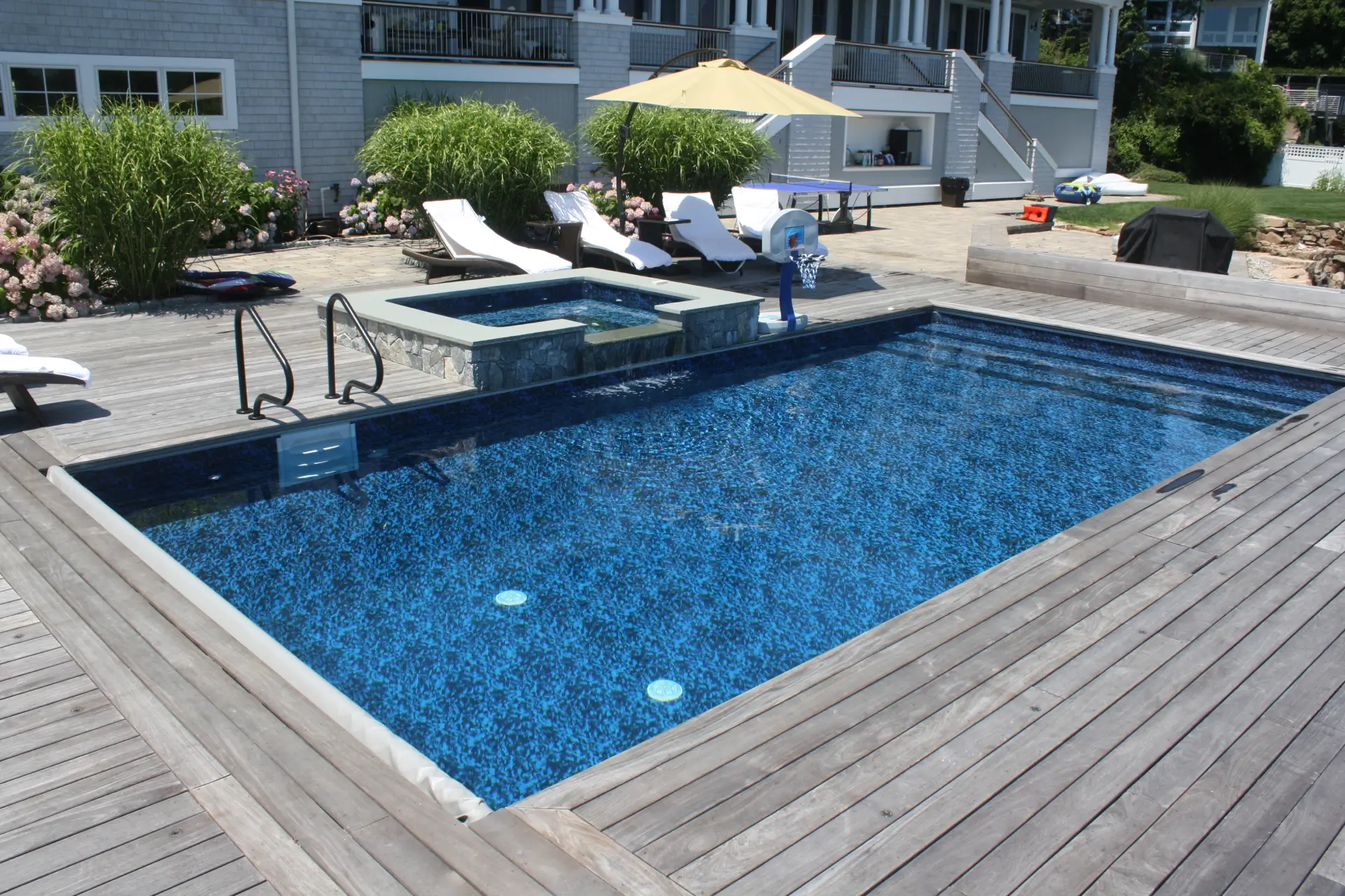 Rectangular in-ground vinyl liner pool with a raised square spa, stone veneer surround, and a spillway waterfall flowing into the pool beside a wood deck with lounge chairs.
