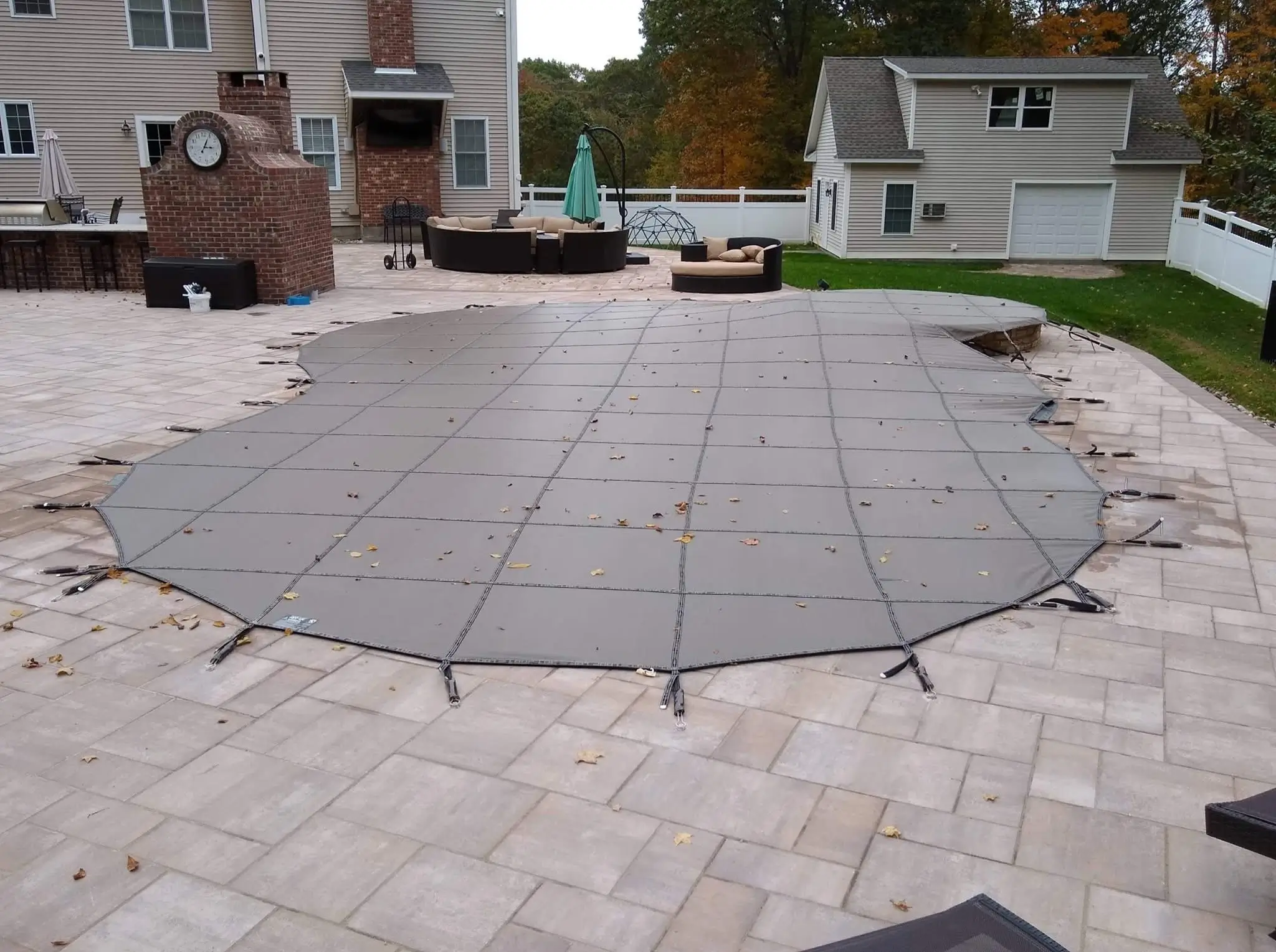 Gray mesh safety cover secured over an in-ground pool on a paved patio behind a house.