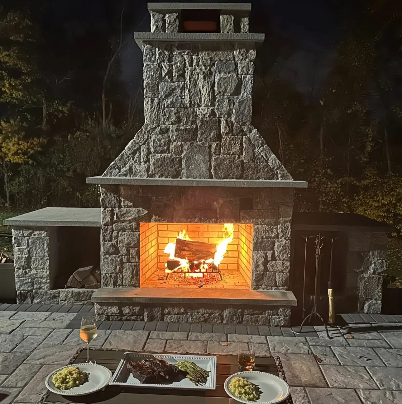 Large stone outdoor fireplace with a burning fire at night, with a small table set for dinner in the foreground.
