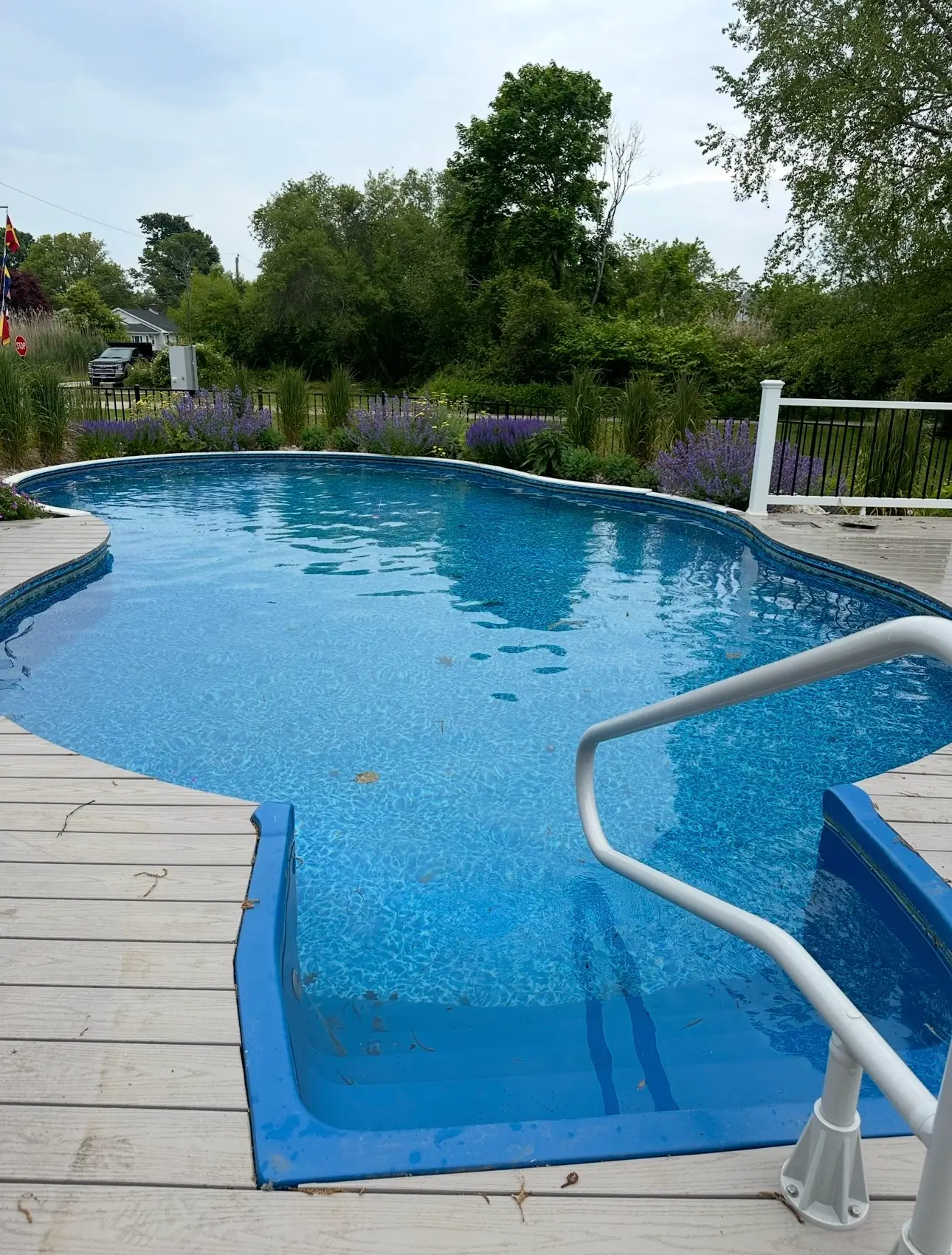 Modern rectangular pool with wide stone decking, a shallow tanning ledge with in-water loungers, and a raised water feature area with seating near a wooded backdrop.