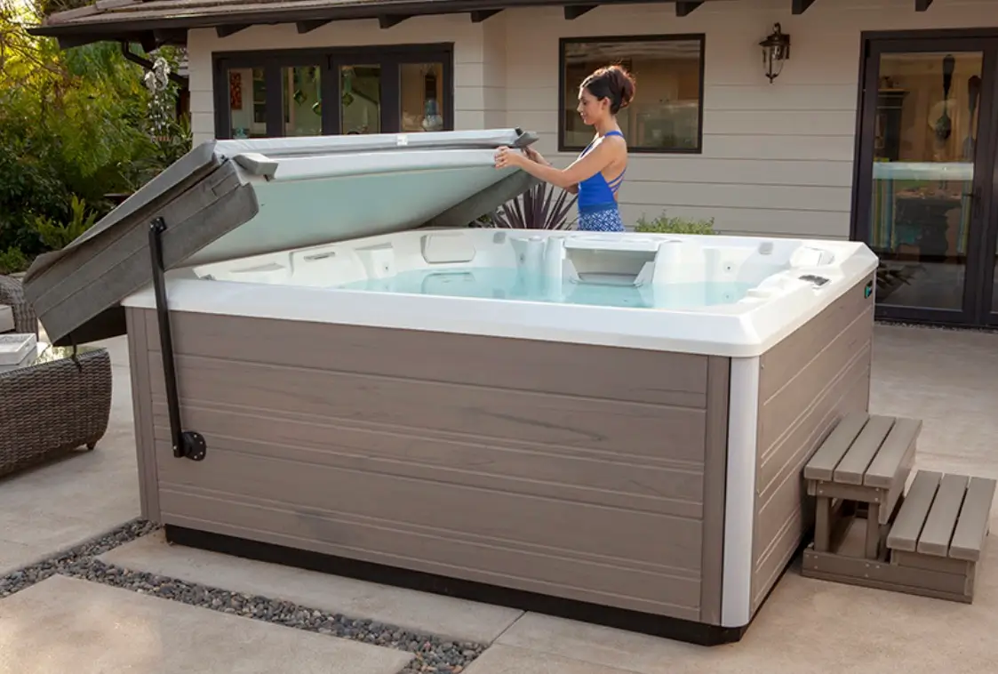 Person lifting the insulated cover on a backyard hot tub beside a house patio.