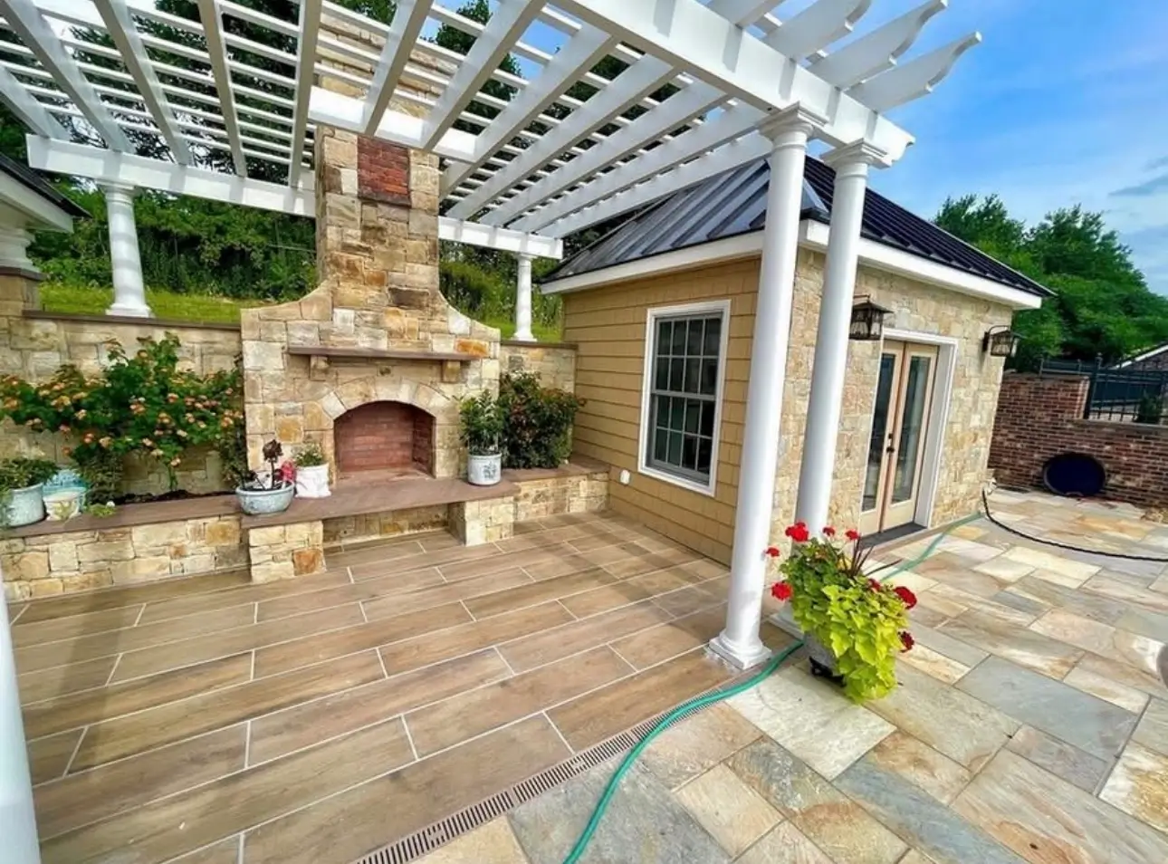 White pergola covering a patio beside a stone outdoor fireplace and a small outbuilding with doors and windows.