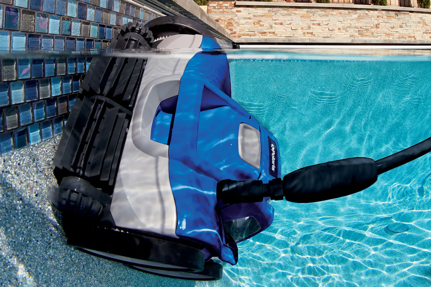 A Polaris robotic pool cleaner with a blue and white body is shown partially submerged, using its brushes to climb a vertically tiled pool wall under clear blue water.