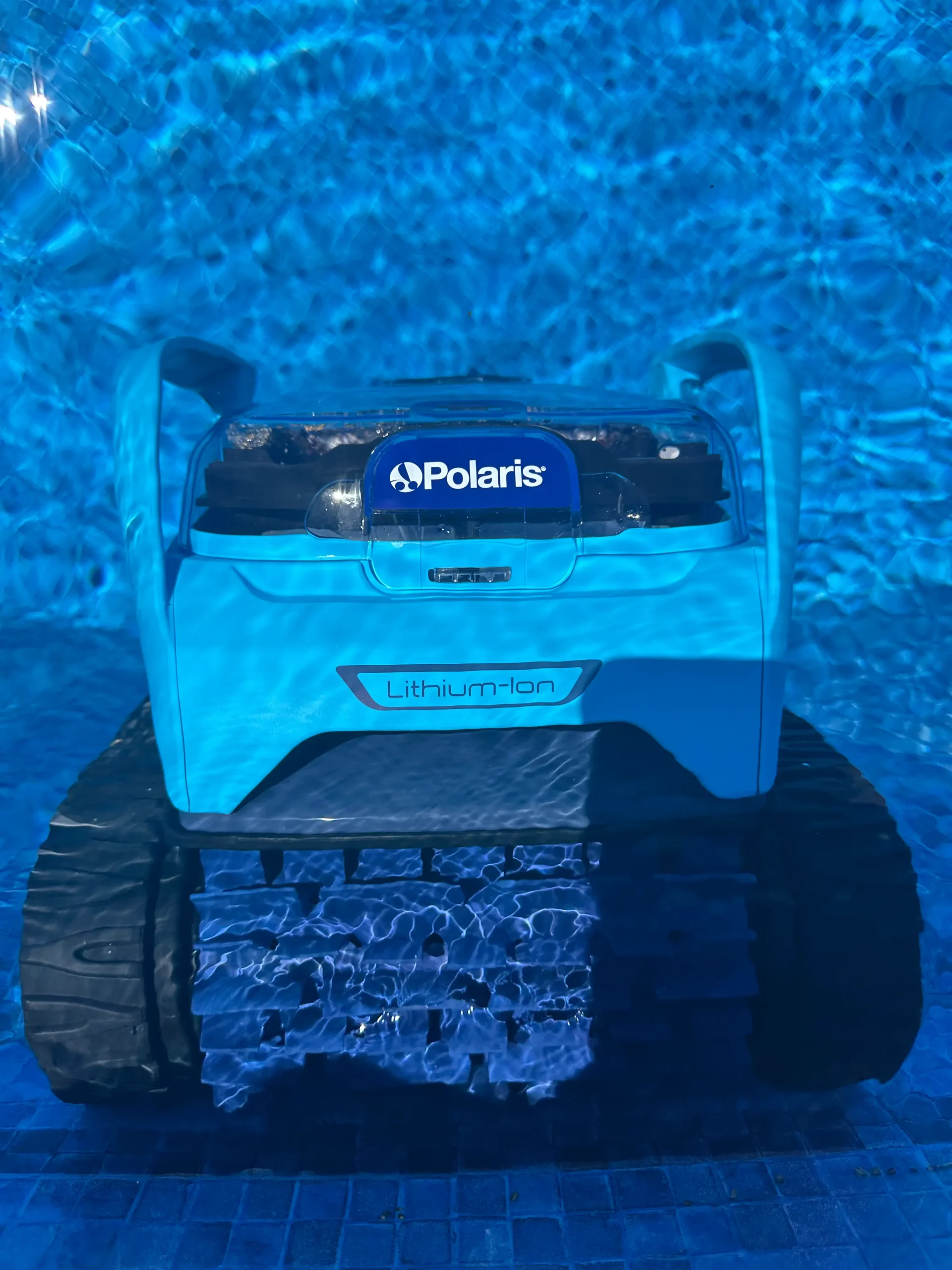 A top-down view of a bright blue Polaris robotic pool cleaner underwater in a swimming pool. The rippling blue water creates a shimmering effect over the device.