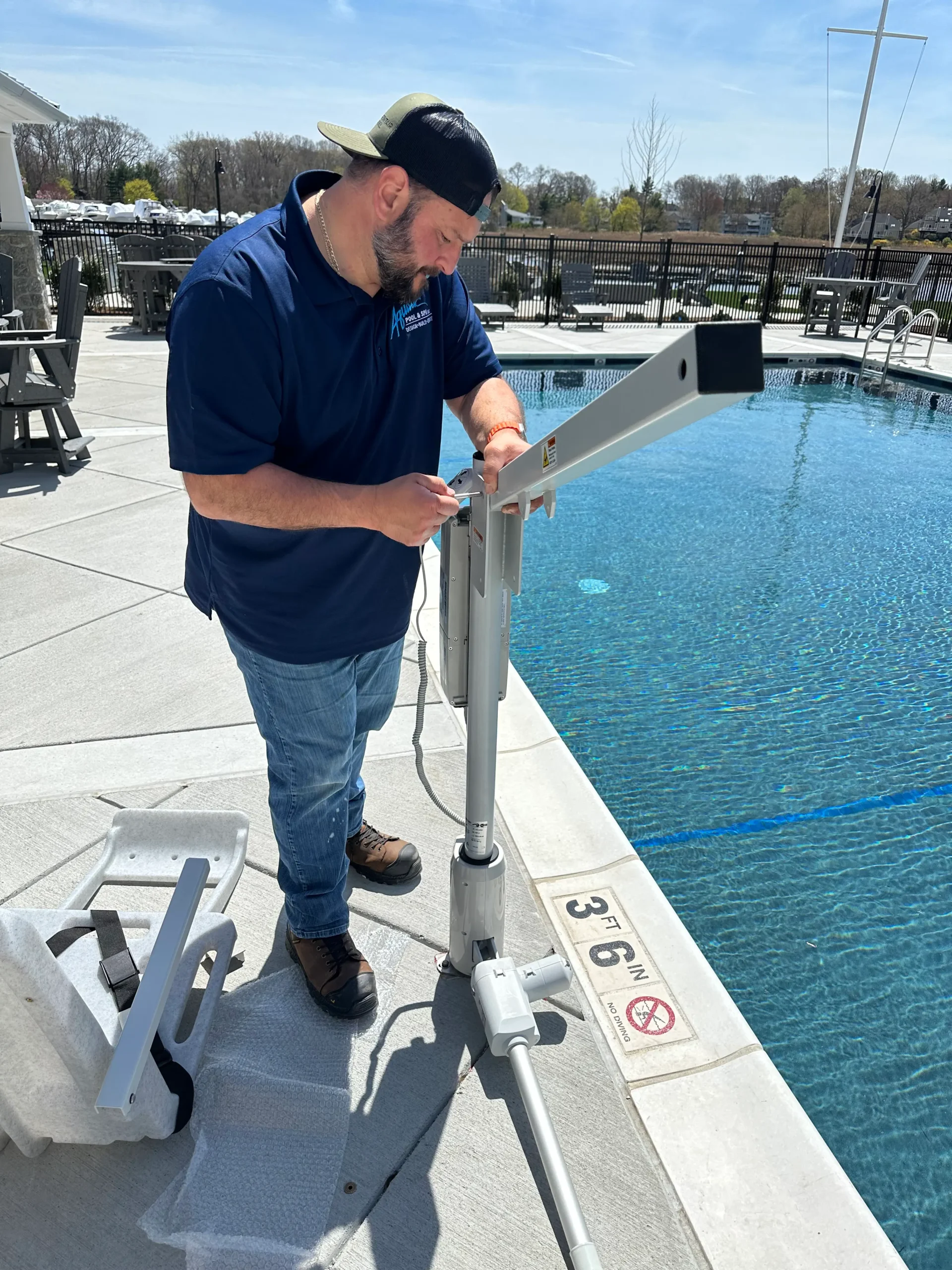 Technician in a navy polo adjusts a pool lift beside a swimming pool on a sunny day.