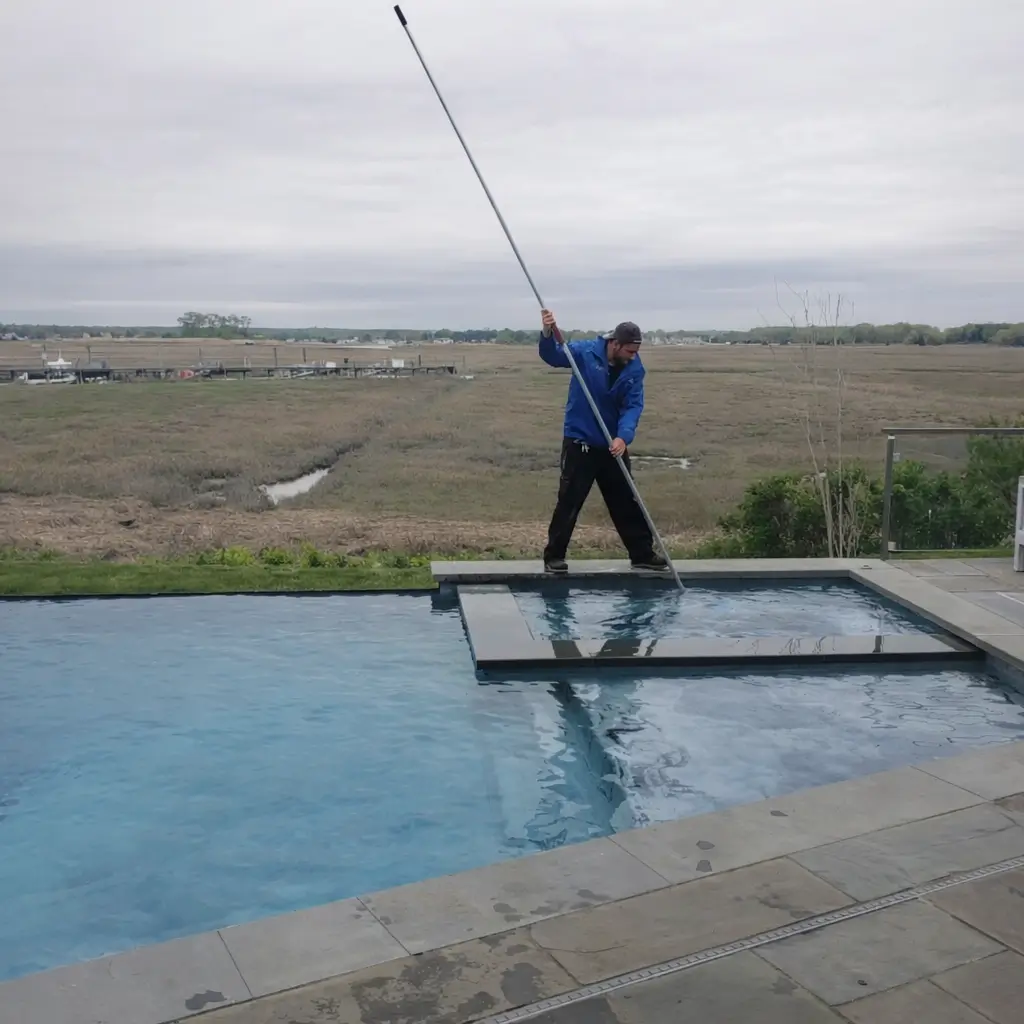 Worker uses a long skimmer pole to clean a pool while standing on the coping, with marshland in the background.