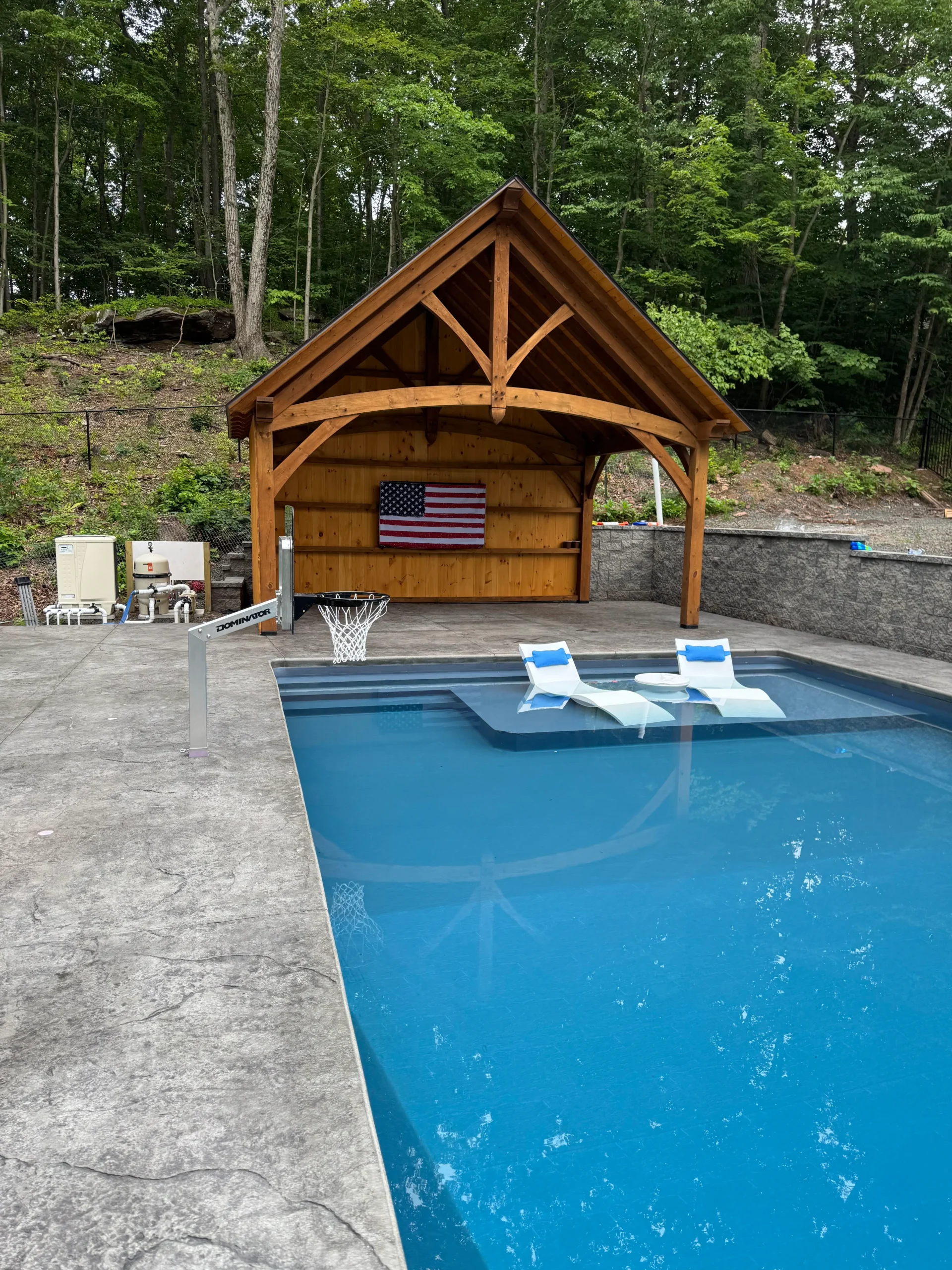 In-ground pool with two in-water loungers, a poolside basketball hoop, and a wooden pavilion set against a wooded backdrop.