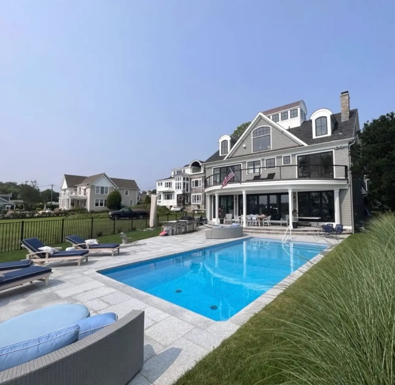 Rectangular pool in front of a large coastal-style home, with blue-cushioned loungers on a stone patio and neighboring houses in the distance.