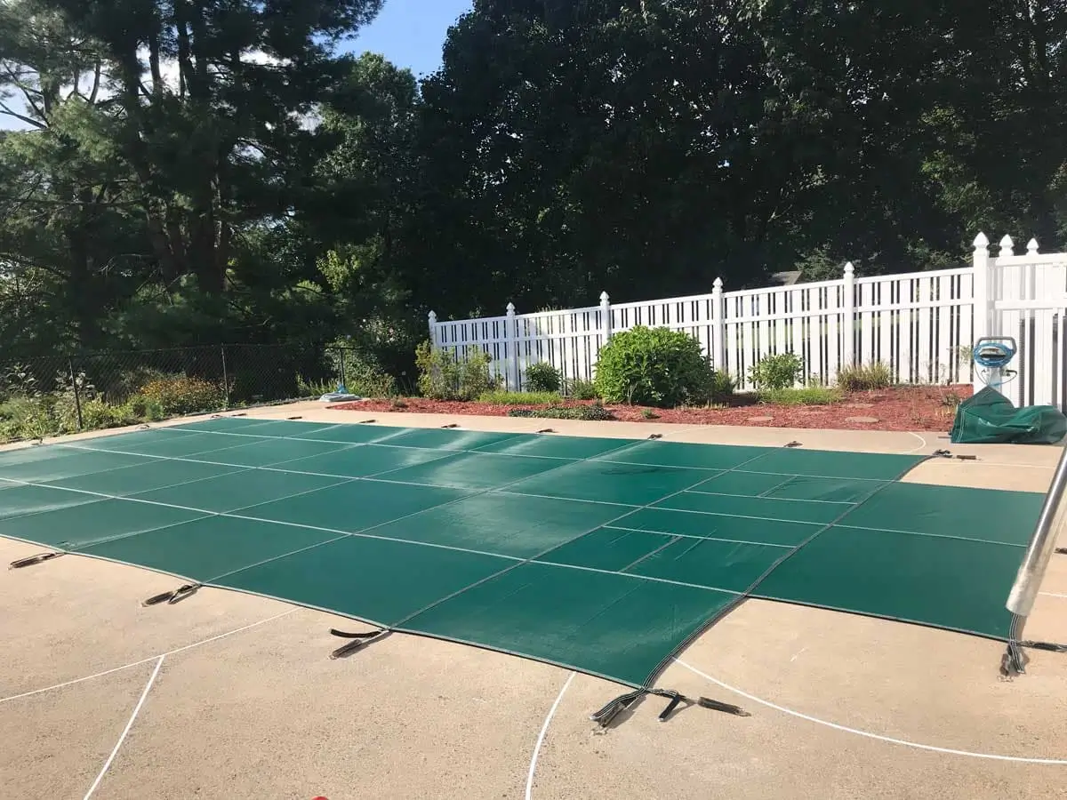 Green pool safety cover stretched and strapped across a pool, with a white fence and trees behind it.