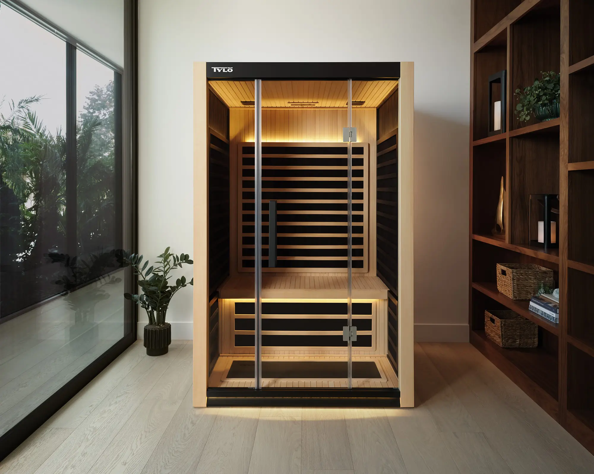A compact Tylö sauna with glass doors and warm interior lighting positioned in a minimalist room between a large window and a tall wooden bookshelf.